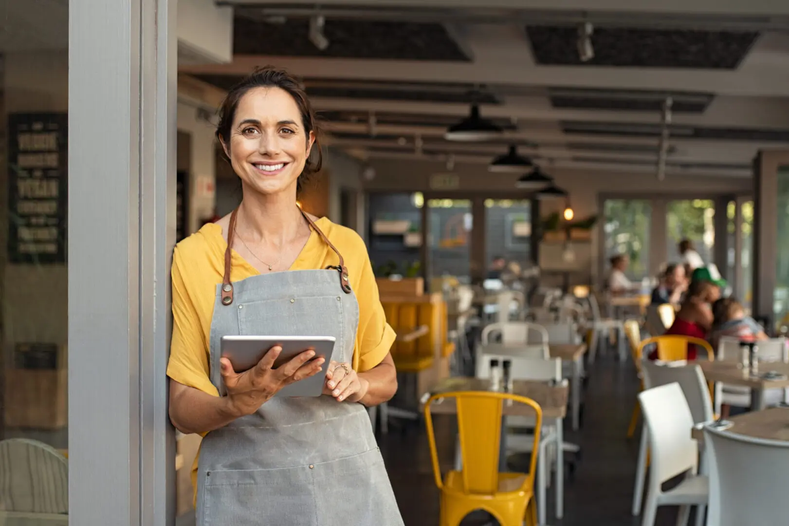 Portrait of happy woman standing at doorway of her store holding digital tablet. Cheerful mature waitress waiting for clients at coffee shop. Successful small business owner in casual clothing and grey apron standing at entrance and looking at camera.